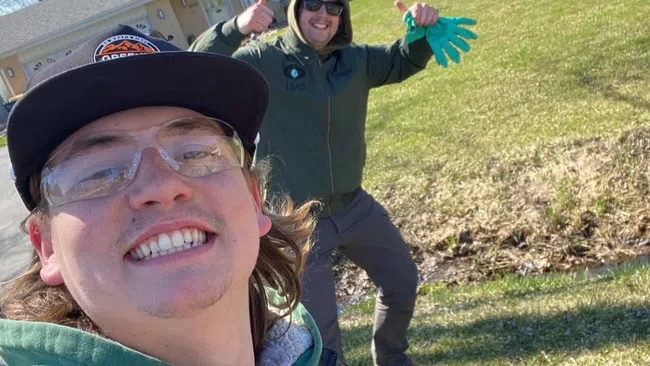 Two men smiling outdoors in a yard with a house in the background on a sunny day.