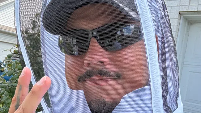 Man wearing a white beekeeping suit, cap, sunglasses, and making a peace sign outdoors.
