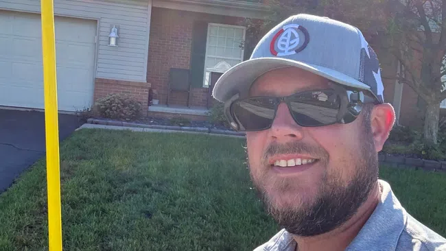 Man wearing sunglasses and a Greenix shirt holds a yellow measuring tool outside a suburban house on a sunny day.