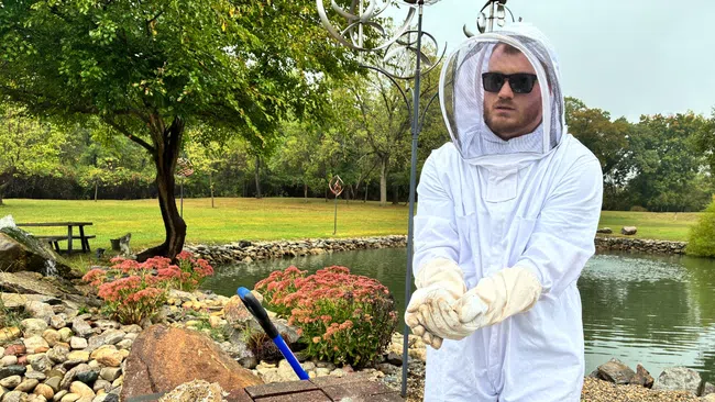 Man in protective beekeeping suit handling honeycomb frames outdoors near pond and garden.