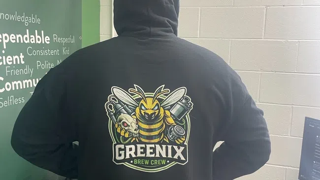 Person wearing black hoodie with Greenix Brew Crew logo featuring a bee holding brewing equipment in an office.