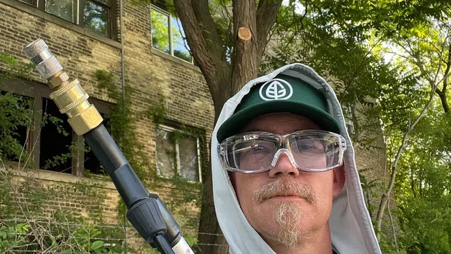 Man in safety glasses and hooded jacket holding a tool outdoors near a brick building and trees.