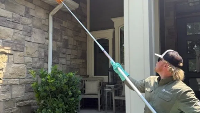 Man in gloves and cap cleaning house gutter using a long brush on a sunny day outside a stone house.