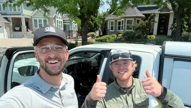 Two men smiling and giving thumbs up next to a white vehicle in a residential neighborhood on a sunny day