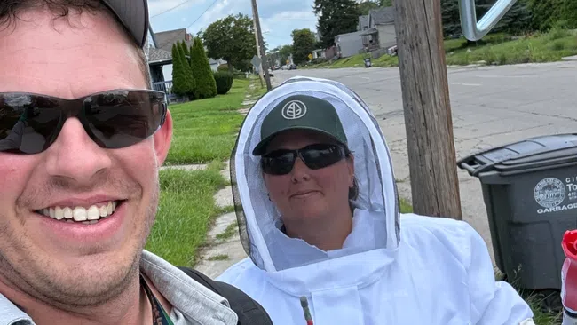 Two people outdoors on a sunny day, one wearing a beekeeping suit and the other smiling with sunglasses and a hat.