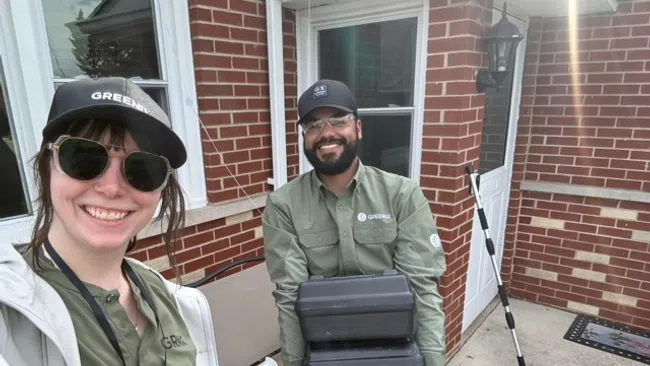 Two smiling pest control workers in green uniforms and gloves standing outside a brick house with equipment.