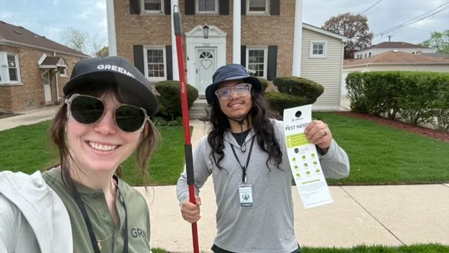 Two pest control workers smiling outside a house, one holding a tool and the other a Pest Nerd flyer.