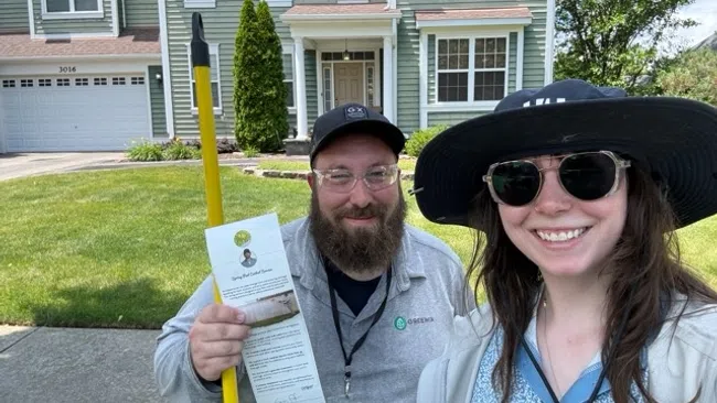 Two smiling lawn care workers standing in front of a residential house holding service flyer and equipment