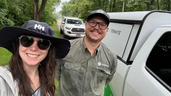 Smiling pest control workers stand by white trucks on a green tree-lined path on a cloudy day