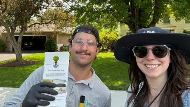 Two people smiling in a sunny yard holding a brochure while wearing gloves and sunglasses, surrounded by green trees and grass.