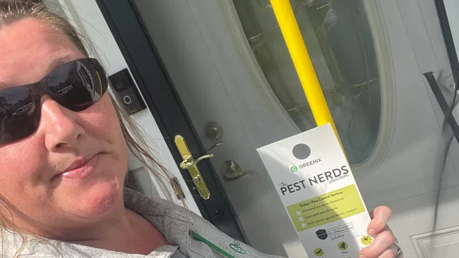Woman in sunglasses holding a pest control door hanger and yellow handle in front of a white door with decorative glass.
