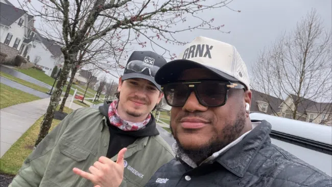Two men outdoors giving thumbs up under a leafless tree on a cloudy day in a suburban neighborhood.
