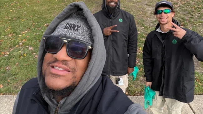 Three smiling men in winter jackets posing on a suburban sidewalk with autumn trees and houses in the background