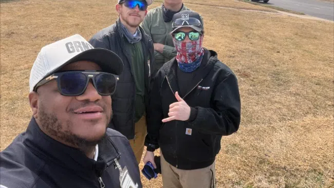 Four men wearing casual outdoor clothing and sunglasses posing for a selfie in a suburban neighborhood with houses and cars.