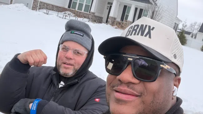 Two men in winter jackets and hats smiling outdoors in front of a snow-covered modern house on a cloudy day