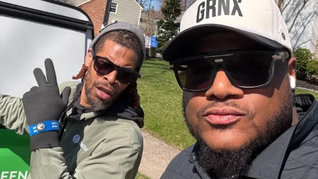 Two men wearing sunglasses outdoors on a sunny day with houses and bare trees in the background.