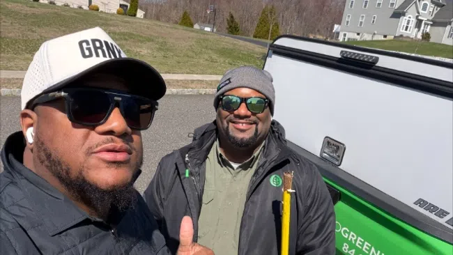 Two pest control workers wearing Greenix jackets pose next to a Greenix company truck in a residential neighborhood.