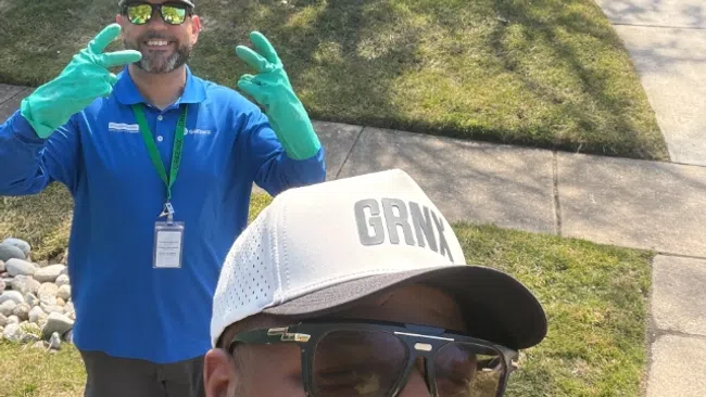 Two men outdoors on a sunny day, one wearing a white GRMX cap and sunglasses, the other in blue with green gloves smiling.