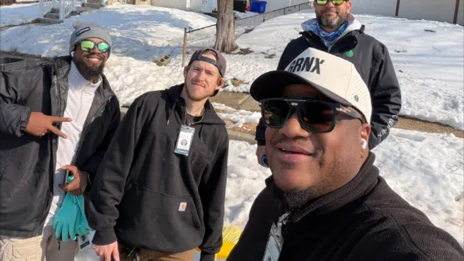 Four men wearing winter clothes and sunglasses pose for a selfie outside in a snowy neighborhood on a sunny day.