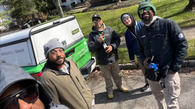 Five pest control workers pose outside a residential home near a white and green service truck on a sunny day.