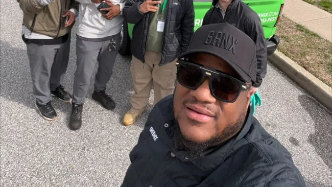 Group of five men posing for a selfie on a suburban street beside green and white work trucks under bare trees.