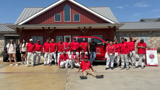 Group of people in red shirts and white pants posing outside a red and gray building with a red truck and signboard on a sunny day