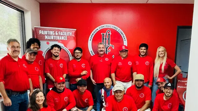Group of employees in red shirts posing inside a Harrison Contracting Company office with branding banners.