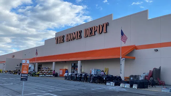 Exterior view of The Home Depot store with orange awnings, outdoor grills, and American flags under a blue sky.
