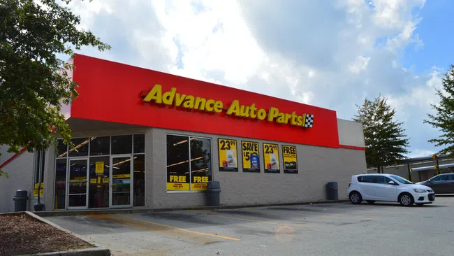 Advance Auto Parts store exterior with red signage, promotional posters, parked cars, and partly cloudy sky.