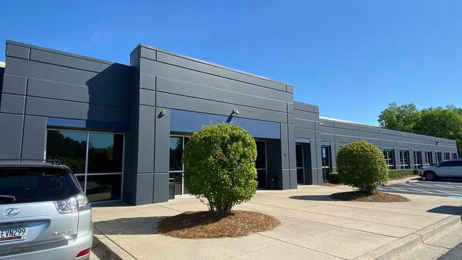 Modern gray commercial building with large windows, two trimmed bushes, and parked cars under clear blue sky.