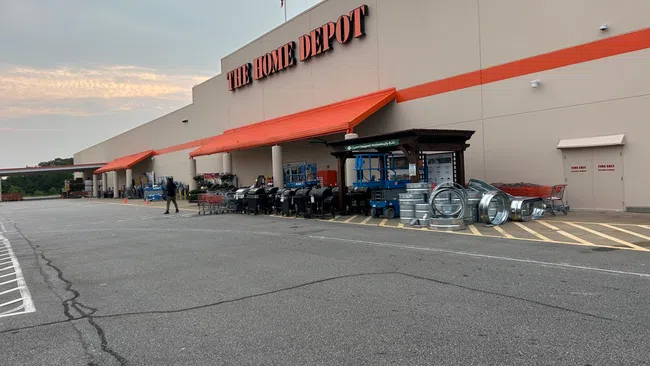 Wide exterior view of The Home Depot with outdoor grills, metal ducts, and shopping carts under overcast sky.