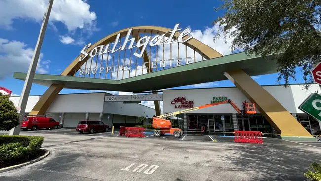 Entrance of Southgate Shopping Center with large arch and storefronts under a blue sky with scattered clouds.