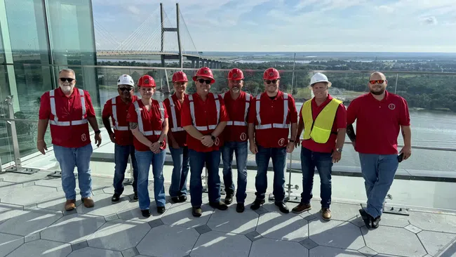 Group of construction workers in red safety vests and helmets standing on rooftop with bridge and river in background