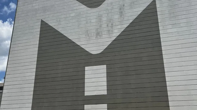 Large gray and white geometric mural resembling the letter M on a building under a partly cloudy sky.