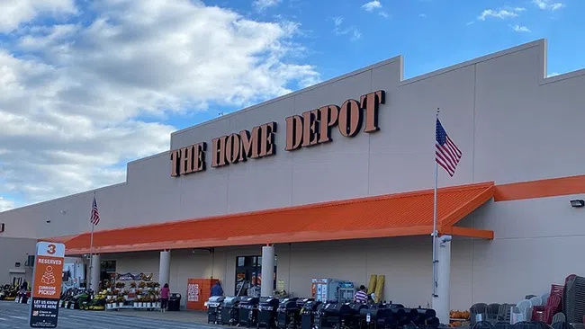 Exterior view of The Home Depot store with orange awning, shopping carts, and American flags under a partly cloudy sky.