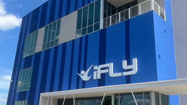 Modern blue iFLY building with large windows and a balcony, under a clear sky with scattered clouds.