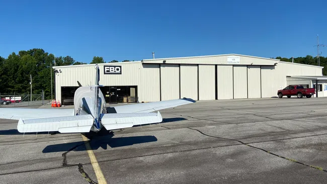 Small private airplane parked on runway in front of white FBO hangar under clear blue sky