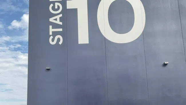 Large dark building labeled Stage 10 with a red van parked in front under a cloudy blue sky