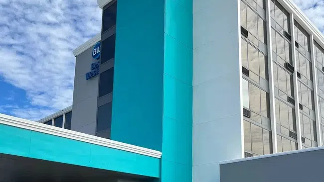 Three people standing outside a modern hotel entrance with teal and white exterior under a blue sky.