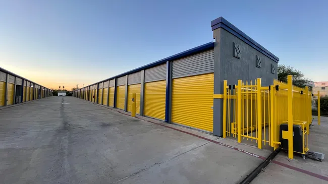 Wide view of a storage facility with yellow roll-up doors and gated entrance under a clear sky at sunset.
