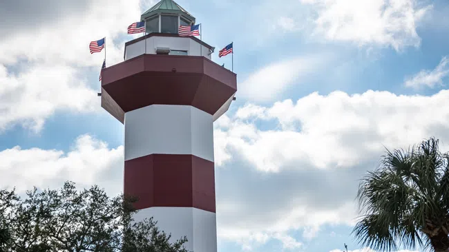 Red and white striped lighthouse with American flags against a partly cloudy blue sky.