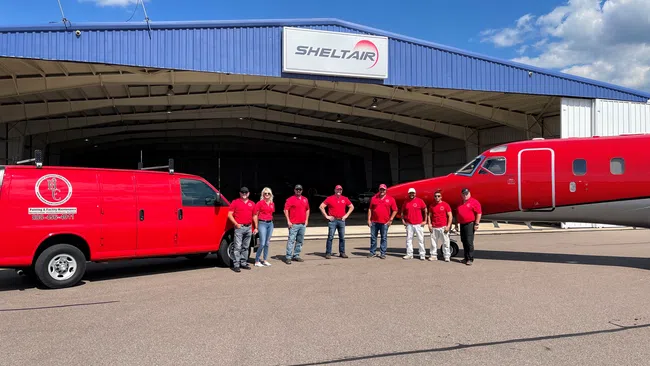 Crew in red shirts stand between red service van and airplane outside SheltaIR hangar under blue sky.