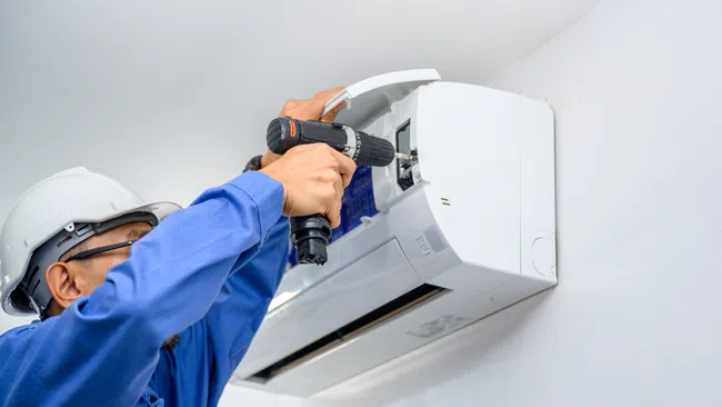 Technician in blue uniform and white helmet installing or repairing a white wall-mounted air conditioning unit with a electric drill.