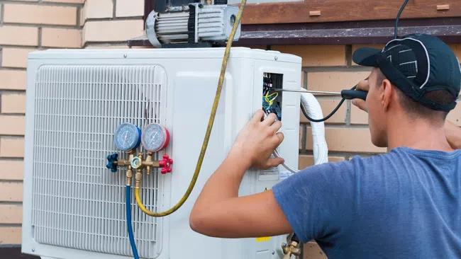 Technician inspecting and repairing an outdoor air conditioning unit with gauges and electrical tools.