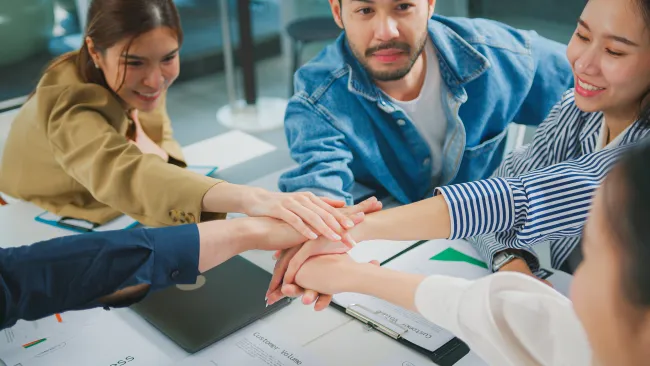 Team of diverse young professionals stacking hands in a collaborative office meeting around documents.
