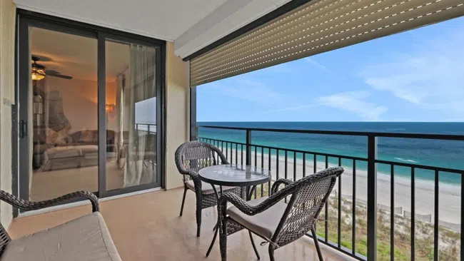 Balcony view with wicker chairs and table overlooking a sandy beach and blue ocean under clear skies.