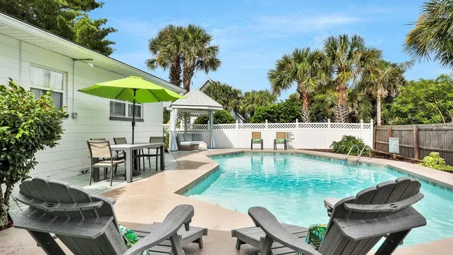 Bright outdoor pool area with lounge chairs, patio table, green umbrella, and palm trees under blue sky
