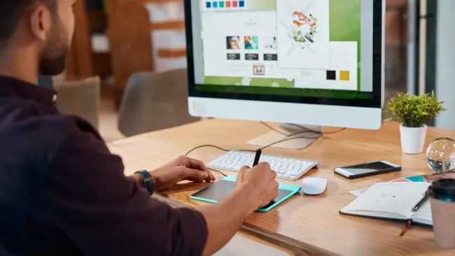 Designer using a digital tablet and stylus while working on a graphic project on a desktop computer screen.