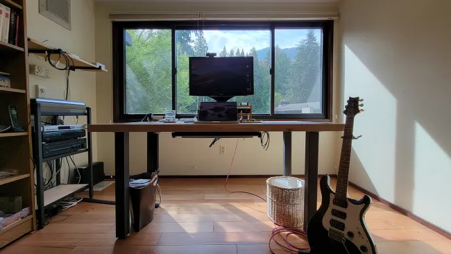 Modern home office with large window, desk, computer monitors, guitar, and light wooden floor under natural daylight.
