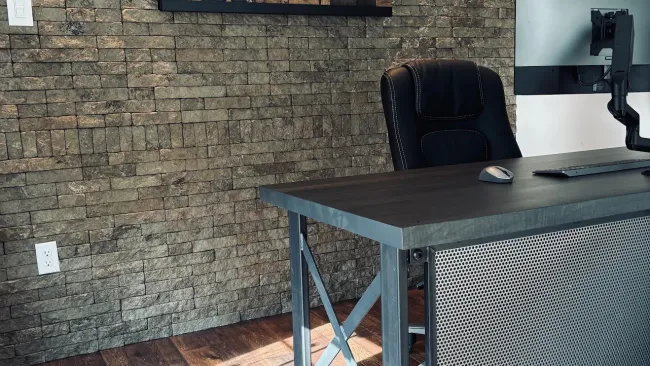 Modern office with stone brick wall, industrial desk, black chair, and wooden American flag decor.